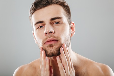 Close up beauty portrait of a handsome bearded man looking at camera with hands on chin isolated over gray backgroundの写真素材