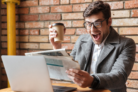 Happy screaming business man in eyeglasses sitting by the table in cafe with laptop computer while reading newspaper and holding cup of coffeeの写真素材