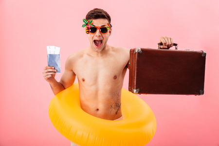 Picture of young happy man standing with rubber ring and sunglasses isolated over pink background. Looking camera holding passport and tickets with suitcase.の写真素材