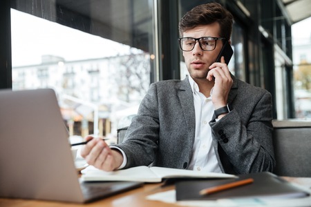 Confused business man in eyeglasses sitting by the table in cafe with laptop computer while talking by smartphone and writing somethingの写真素材