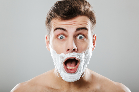 Close up portrait of a shocked man with shaving foam on his face isolated over gray backgroundの写真素材