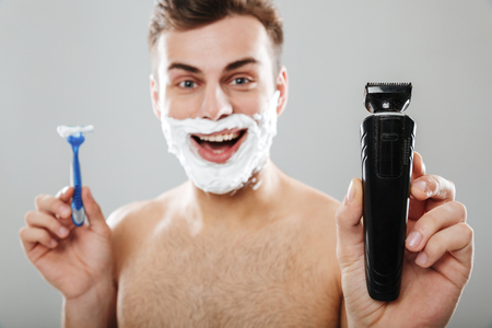Close up portrait of a cheerful man with shaving foam on his face holding a razor and a trimmer isolated over gray backgroundの写真素材