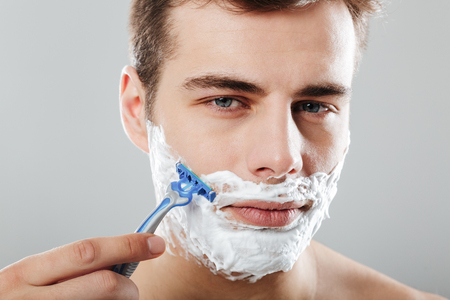 Close up portrait of a serious man with shaving foam on his face shaving with a razor isolated over gray backgroundの写真素材