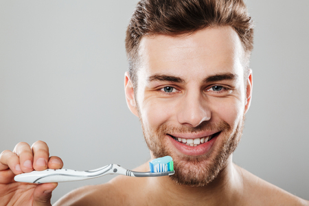 Close up portrait of a smiling half man holding toothbrush with toothpaste isolated over gray backgroundの写真素材