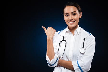 Young happy lady with stethoscope looking camera and pointing at copy space isolated over blackの写真素材