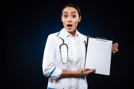 Shocked lady doctor in white medical gown showing blank folder isolated over blackの写真素材