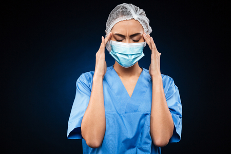 Tired female doctor in medical mask and cap has a headache isolated over blackの写真素材