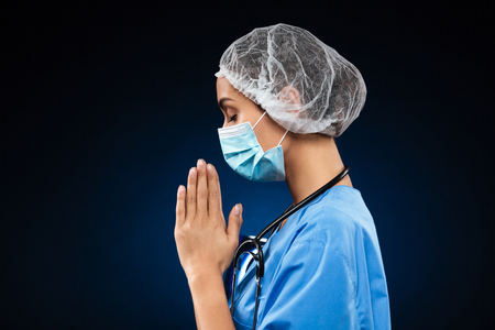 Side view of praying doctor in uniform and medical mask and cap isolated over blackの写真素材