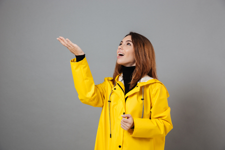 Portrait of an excited girl dressed in raincoat and rubber boots posing while standing with outstretched hand isolated over gray backgroundの写真素材