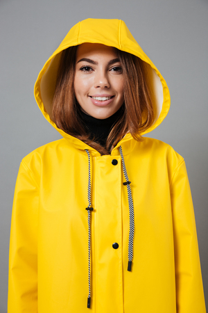 Portrait of a cheerful girl dressed in raincoat posing with hood on her head and looking at camera isolated over gray backgroundの写真素材