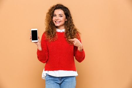 Portrait of a smiling young girl pointing finger at blank screen mobile phone isolated over beige backgroundの写真素材