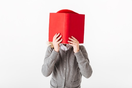 Close up portrait of a little schoolgirl dressed in uniform covering face with a book isolated over white backgroundの写真素材