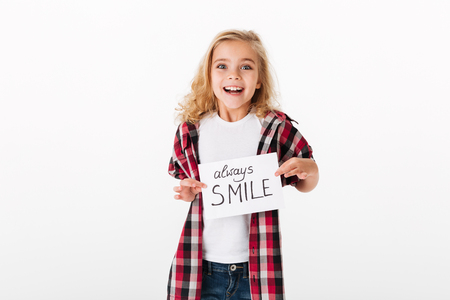 Portrait of an excited little girl holding sheet of paper with an inscription Always smile isolated over white backgroundの写真素材