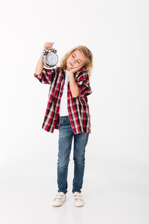 Full length portrait of a sleepy little girl holding alarm clock while standing isolated over white backgroundの写真素材