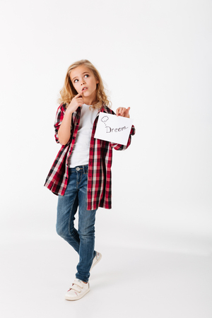 Full length portrait of a wondering little girl holding sheet of paper with an inscription Dream isolated over white backgroundの写真素材