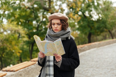 Portrait of a concentrated girl dressed in autumn clothes holding city guide map while walking...