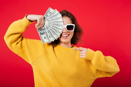 Portrait of a smiling woman in sunglasses posing while holding bunch of money banknotes isolated...