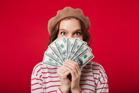 Portrait of a young woman wearing beret holding bunch of money banknotes at her face isolated over pink backgroundの写真素材