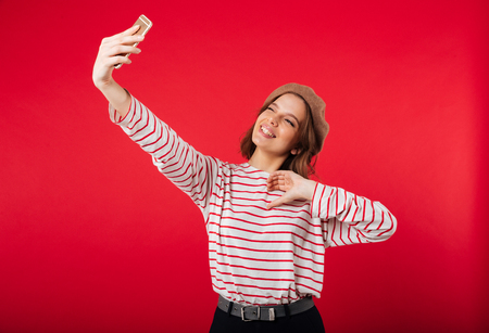 Portrait of a lovely woman wearing beret taking a selfie isolated over pink backgroundの写真素材
