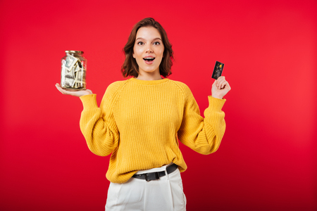 Portrait of a happy woman holding jar full of money and a credit card banknotes isolated over pink backgroundの写真素材
