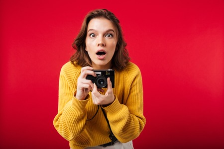 Portrait of a shocked woman holding vintage camera isolated over pink backgroundの写真素材