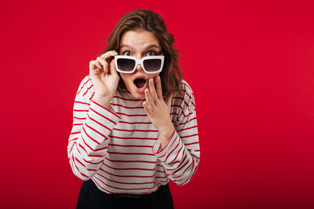 Portrait of a shocked woman in sunglasses posing while standing and looking at camera isolated over pink backgroundの写真素材