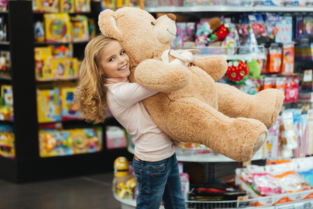 Smiling little girl holding big teddy bear while standing at the supermarketの写真素材