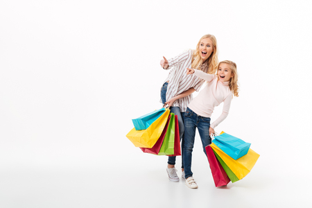 Full length portrait of a shocked mother and her little daughter standing with shopping bags and pointing fingers away isolated over white background