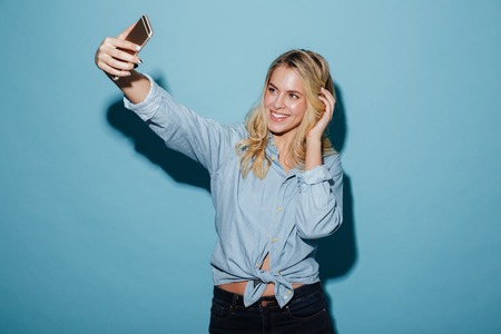 Cheerful blonde woman in shirt making selfie on smartphone over blue background