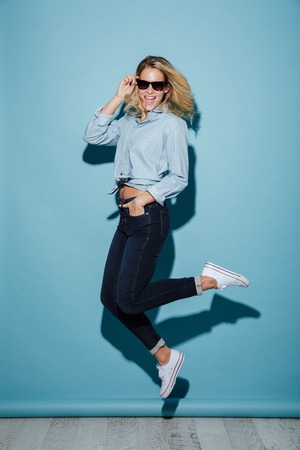 Full length image of Happy blonde woman in shirt and sunglasses jumping with arm in pocket and...
