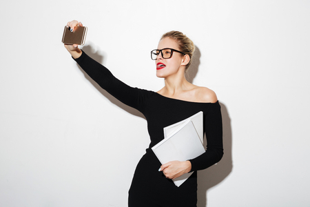 Cheerful business woman in dress and eyeglasses making selfie on smartphone while holding documents over gray backgroundの写真素材