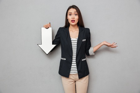 Surprised asian business woman shrugs her shoulders and pointing with paper arrow down while looking...
