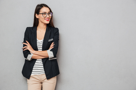 Smiling asian business woman in eyeglasses with crossed arms looking away over gray backgroundの写真素材