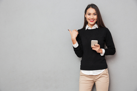 Smiling asian woman in business clothes holding smartphone and pointing on copyspace while looking at the camera over gray background