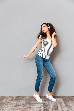 Full size view of charming young woman in striped t-shirt and jeans dancing, while listening melodies via headphones over grey backgroundの写真素材