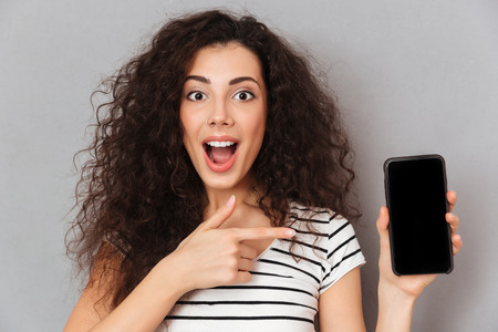 Close up photo of optimistic woman with ring in nose pointing index finger like advertising her smartphone, meaning good choice against grey wallの写真素材