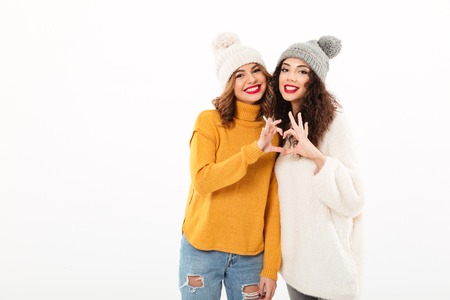Two smiling girls in sweaters and hats making heart sign while looking at the camera over white backgroundの写真素材