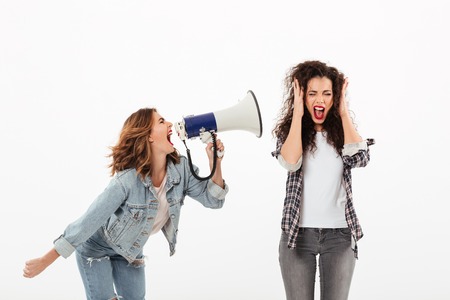 Confused curly woman covering her ears while second girl screaming at her with megaphone over white backgroundの写真素材