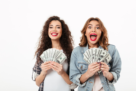 Two happy girls holding money and rejoices while looking at the camera over white backgroundの写真素材
