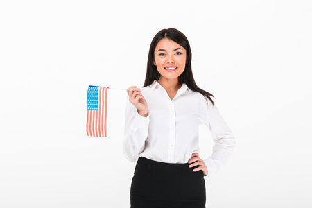 Portrait of a smiling asian businesswoman holding american flag while standing and looking at camera isolated over white backgroundの写真素材