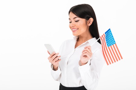 Portrait of a satisfied asian businesswoman holding american flag while standing and using mobile phone isolated over white backgroundの写真素材