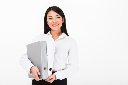Portrait of a smiling asian businesswoman holding binders and looking at camera isolated over white backgroundの写真素材