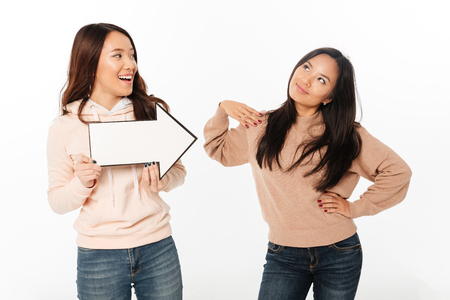 Photo of two asian positive ladies sisters standing isolated over white background holding arrow.の写真素材