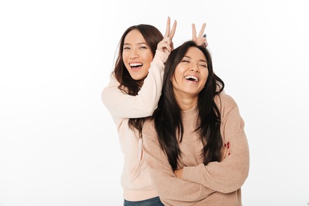 Picture of two asian cute ladies sisters standing isolated over white background looking camera have fun.