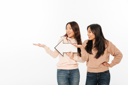 Image of two asian positive ladies sisters standing isolated over white background holding arrow showing copyspace.の写真素材