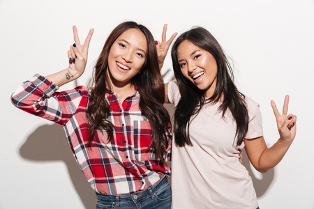 Image of two asian pretty cheerful ladies sisters hugging with each other. Looking camera showing peace gesture.の写真素材
