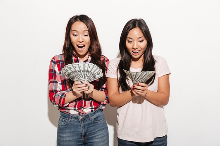 Photo of two asian pretty shocked cute ladies sisters standing isolated over white background. Looking aside holding money.の写真素材