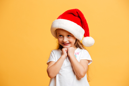 Picture of little cheerful girl standing isolated over yellow background wearing christmas santa hat. Looking aside.の写真素材