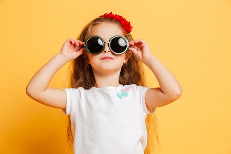 Photo of little cute serious pretty girl standing isolated over yellow background wearing sunglasses looking camera.の写真素材