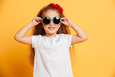 Photo of little cute pretty girl standing isolated over yellow background wearing sunglasses looking camera.の写真素材